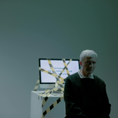 A senior adult sits indoors next to a computer wrapped in caution tape, symbolizing job loss and unemployment.